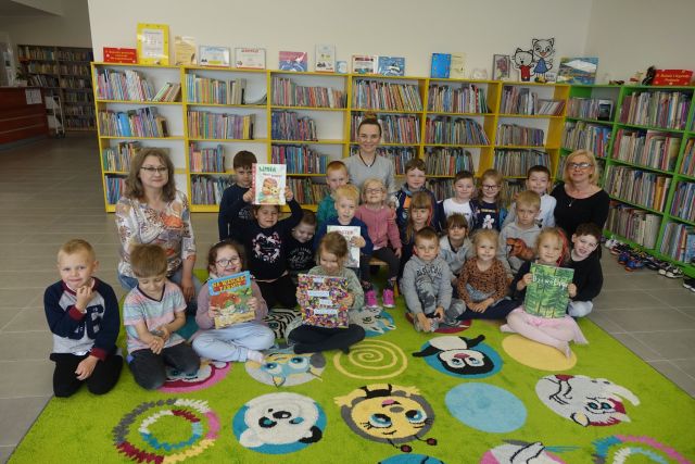 na zielonym dywanie siedzą dzieci i trzy kobiety, za nimi żółte regały z książkami