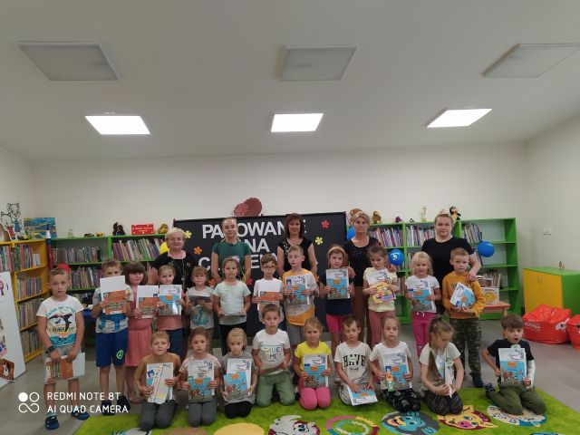 w pierwszym rzędzie dzieci siedzą na zielonym dywanie, na nimi stoją dzieci, za nimi 4 kobiety, za n imi czarna tablica