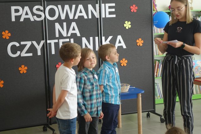 4 chłopców stoi przed kobietą trzymającą w dłoniach kartkę, za n imi czarna tablica