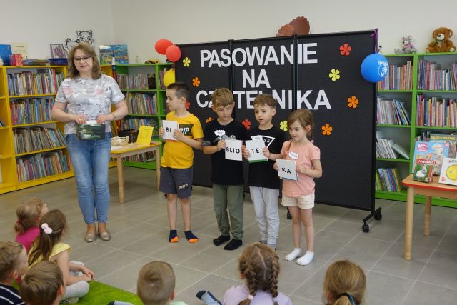 3 chłopców i dziewczynka stoją i trzymają w dłoniach kartki z literami, z lewej strony stoi kobieta, za nimi czarna tablica i zielone regały z książkami