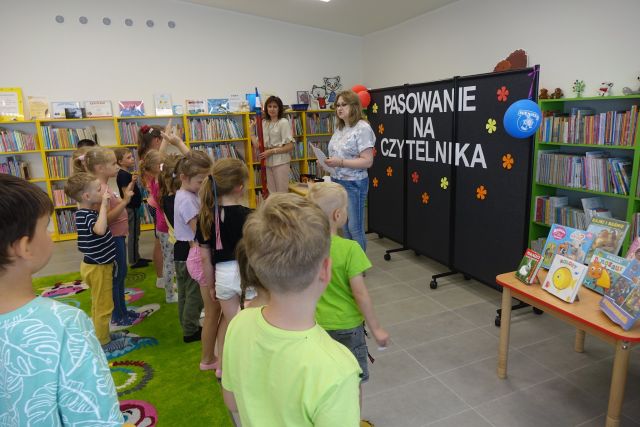 tyłem stoją dzieci, przed nimi dwie kobiety, jedna trzyma w dłoniach ogromny czerwony ołówek, za nimi czarna teblica, z lewej strony żółte regały z książkami