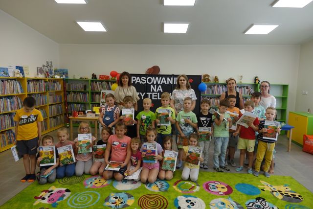 w pierwszym rzędzie na zielonym dywanie siedzą dzieci, za nimi stoją dzieci i za nimi 4 kobiety, dzieci trzymają w dłoniach dyplomy i książki