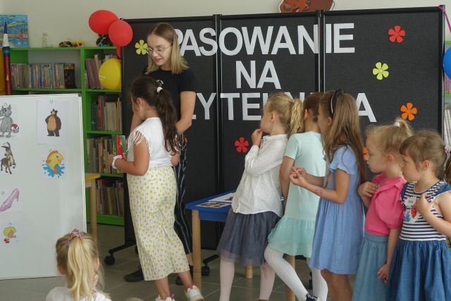 dzieci stoją przed białą tablicą z ilustracjami z bajek, za nimi stoi kobieta, za nią czarna tablica