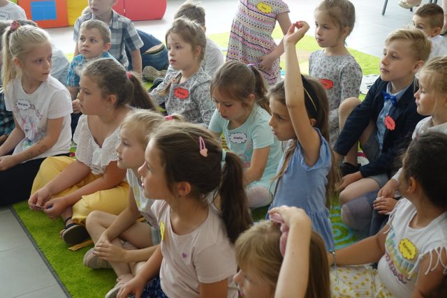 dzieci siedzą na zielonym dywanie, niektóre maja podniesioną do góry rękę