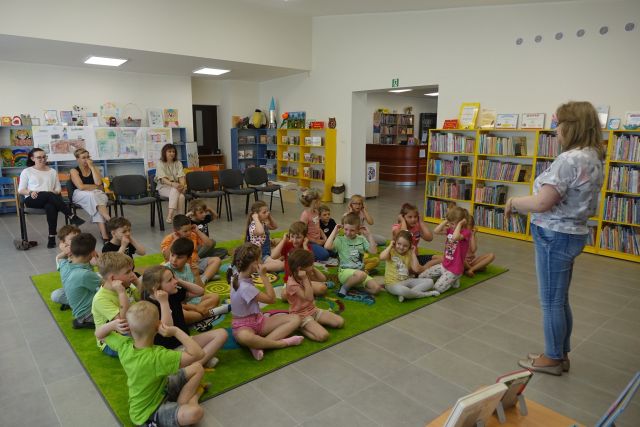 na zielonym dywanie siedzą dzieci, za nimi na krzesłach 3 kobiety, za nimi żółte i niebieskie regały z książkami, przed dziećmi stoi kobieta