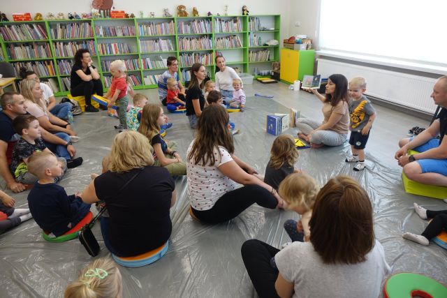 na siwej podłodze przykrytej folią siedzą dzieci z mamami, tatami i babciami, przed nimi siedzi kobieta