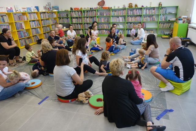 na siwej podłodze przykrytej folią siedzą dzieci z mamami, tatami i babciami, przed nimi siedzi kobieta