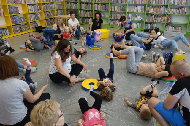 na siwej podłodze przykrytej folią siedzą dzieci z mamami, tatami i babciami, przed nimi siedzi kobieta z nogami do góry uniesionymi