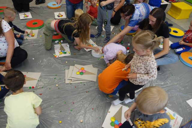 na siwej podłodze przykrytej folią dzieci i ich opiekunowie kleją kulki na kartkach z narysowanymi lodami