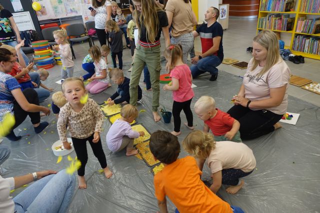 na siwej podłodze przykrytej folią dzieci z opikunami bawią się w miskach z różnymi materiałami, dziewczynka dmuch w żółte piórko