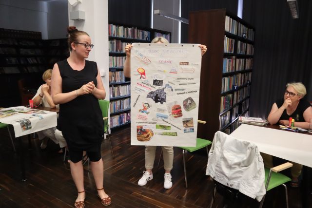 w tle regały z książkami, przed nimi kobieta trzymająca duży arkusz papieru z przyklejonymi wycinkami z gazet, druga kobie stoi z lewej strony patrząca na papier