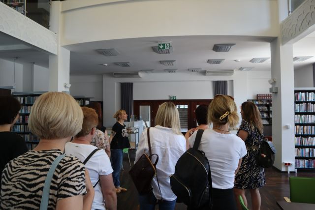 kobiety stoją tyłem, przed nimi kobieta opowiadająca na tle białej ściany