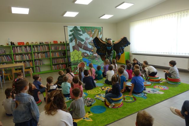 na zielonym dywanie na okrągłych płaskich kolorowych pufach siedzą dzieci, przed nimi stoi kobieta z rozłożonymi rękoma ubranymi w pióra, za nią plakat żubra pompika i zielone regały z książkami