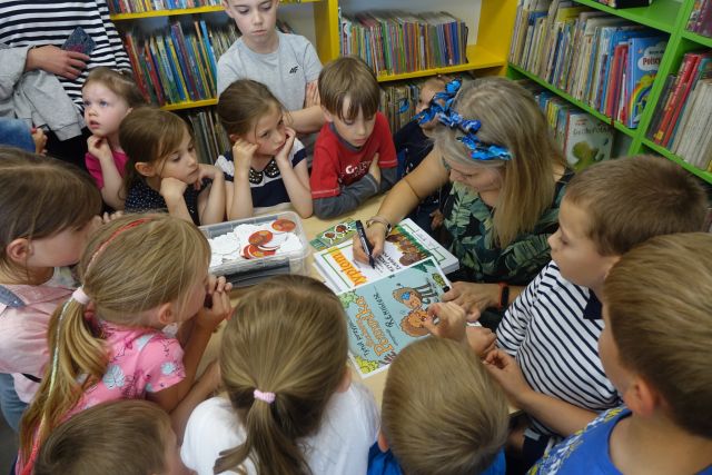 przy brązowym stole siedzi kobieta w niebieskiej opasce i podpisuje dyplomy, wokół stołu stoją dzieci