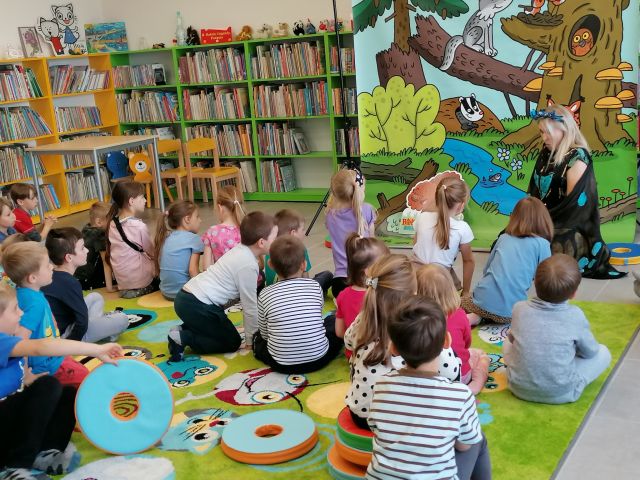 na zielonym dywanie na okrągłych płaskich kolorowych pufach siedzą dzieci, przed nimi siedzi kobieta, za nią plakat żubra pompika i zielone regały z książkami