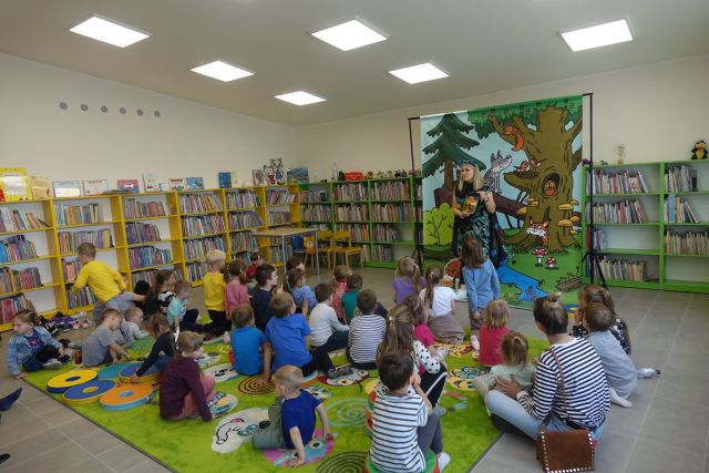 na zielonym dywanie na okrągłych płaskich kolorowych pufach siedzą dzieci, przed nimi stoi kobieta, za nią plakat żubra pompika i zielone regały z książkami