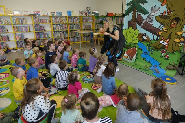 na zielonym dywanie na okrągłych płaskich kolorowych pufach siedzą dzieci, przed nimi stoi kobieta z rękoma ubranymi w pióra, za nią plakat żubra pompika i zielone regały z książkami