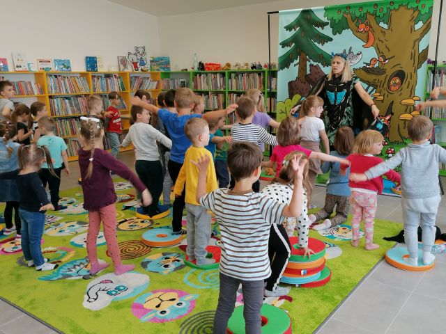 na zielonym dywanie stoją dzieci i ćwiczą dzieci, przed nimi stoi kobieta z rozłożonymi rękoma ubranymi w pióra, za nią plakat żubra pompika i zielone regały z książkami
