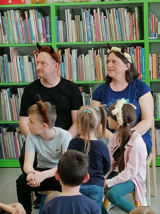 na krzesłach siedzą z przodu chłopiec i dziewczynka, z tyłu kobieta i mężczyzna, za nimi zielone regały z książkami