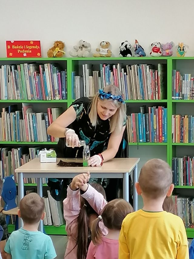 na zielonym dywanie na okrągłych płaskich kolorowych pufach siedzą dzieci, przed nimi stoi kobieta robiąca doświadczenie pochylona nad brązowym stołem za nią plakat żubra pompika i zielone regały z książkami