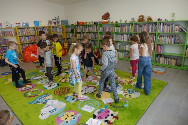 po zielonym dywanie chodzą w kółko dzieci, za nimi zielone i żółte regały z książkami