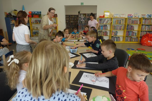 przy brązowych stołach siedzą dzieci, nad nimi stoją rodzice. Dzieci malują na kartkach