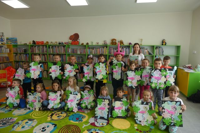 na zielonym dywanie siedzą dzieci, za nimi stoi rzą dzieci i dorosłą kobieta, za nimi zielone regały z książkami. Wszyscy trzymają w dłoniach kartki z rysunkami flaminga