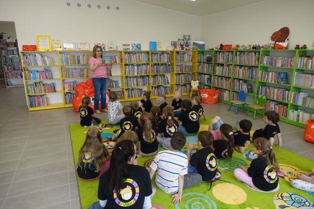 na zielonym dywanie siedzą tyłem dzieci, przed nimi stoi kobieta, za nią żółte regały z książkami