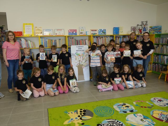 przed żółtymi regałami Z książkami stoją dzieci i 3 osoby dorosłe, w środku pomiędzy nimi stoi plakat z drzewem. Dzieci mają w dłoniach kartki