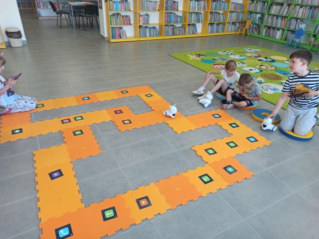 na siwej podłodze leżą pomarańczowe duże puzle, przed  nimi siedzą dzieci i bawią się białymi robotami