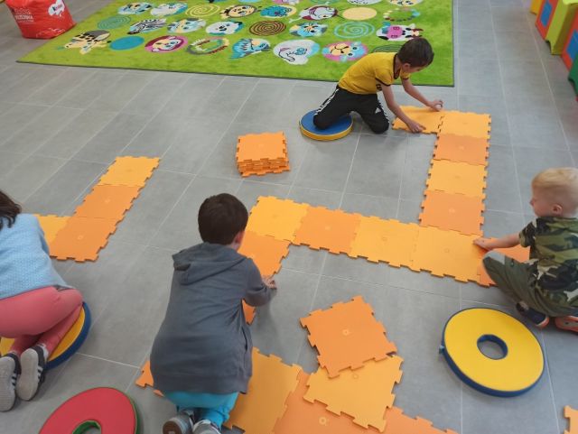 na siwej podłodze siedzą trzej chłopcy i układają pomarańczowe duże puzle, za nimi zielony dywan