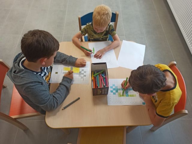 przy brązowym stole siedzą trzej chłopcy i rysują na białych kartkach