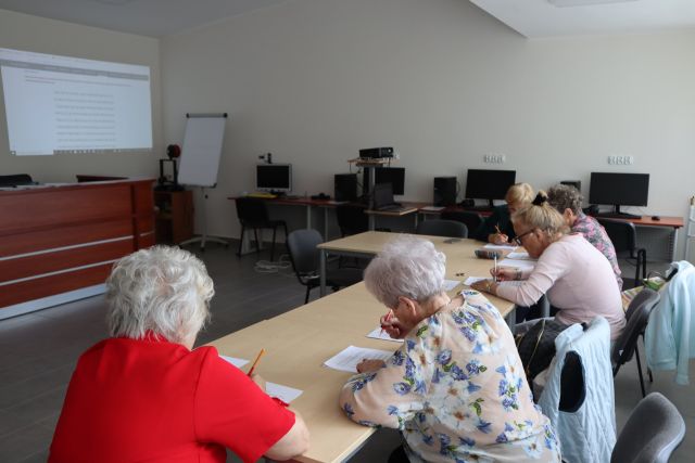 przy brązowych stołach siedzą starsze kobiety i piszą ma kartkach