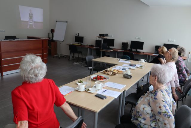 przy brązowych stołach siedzą starsze kobiety z rękoma do tyłu, na stołąch leżą kartki i poczęstunek