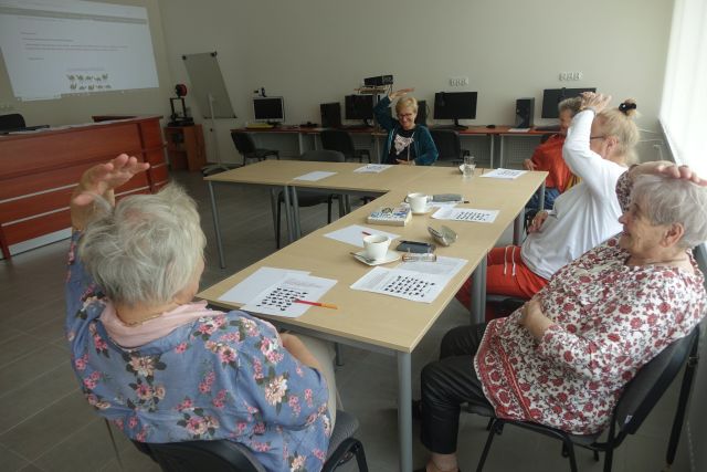 przy brązowych stołąch siedzi 5 starszych kobiet, trzymają ręce na głowami