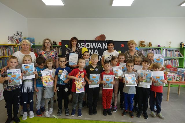 w dwóch rzędach stoją dzieci, w rękach trzymają dyplomy i książki, za nimi stoją 4 dorosłe kobiety, za nimi czarna tablica z napisem pasowanie na czytelnika