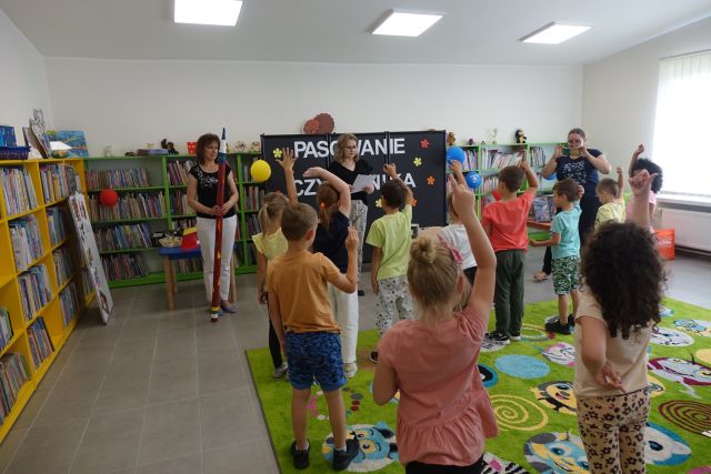 na zielonym dywanie stojątyłęm dzieci, przed nimi dwie kobiety, jedna ma wdłoniach ogromny czerwony ołówek