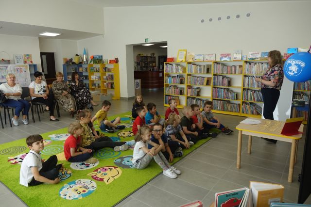 na zielonym dywanie siedzą dzieci, z ich lewej strony żółte regały z książkami