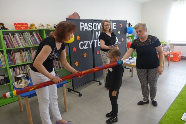 kobeta w czarnej bluzce i białych spodniach dotyka dyżym czerwonym ołówkiem ramię dziecka, za dzieckiem stoi kobieta, w tle kobieta, a za nią czarna tablica