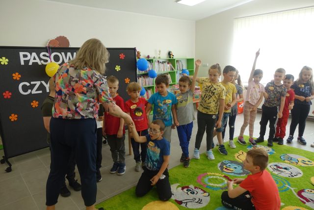 z lewej strony stoi kobieta, przed nią stoją dzieci, za nimi czarna tablica