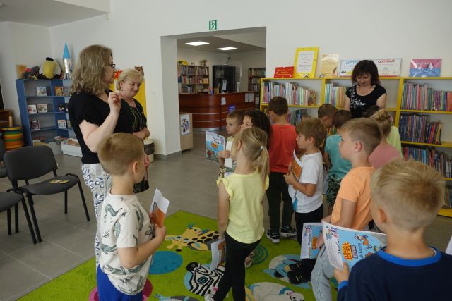 dzieci stoją na zielonym dywanie, przed nimi dwie kobiety i żółte regały z książkami