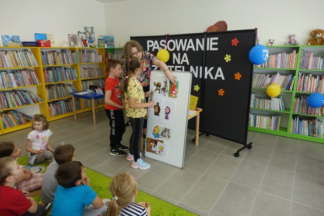 na siwej podłodze przed białą tablicą stoji swójka dzieci i kobieta, któa wpina do tablicy ilustracje postaci z bajek