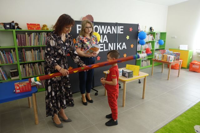 z lewej strony stoi kobieta i trzyma w dłoniach dużą czerwoną kredkę, obok niej stoi kobieta, przed nią chłopiec z ołówkeim położonym na jego ramieniu