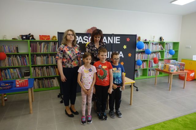 jedna dziewczynka i dwóch chłopców stoją, za nimi dwie kobiety na tle czarnej tablicy