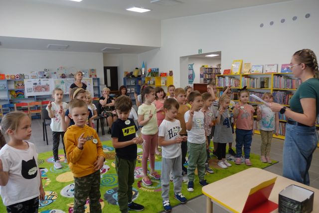 dzieci stoją na zielonym dywanie, za nimi 3 kobiety, w tle niebieskie i żółte regały