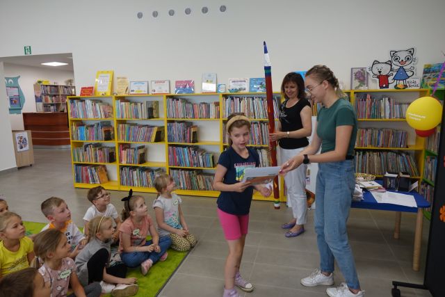 kobieta wręcza dziewczynce dyplom i książkę, z lewej strony na zielonym dywanie siedzą dzieci, z tyłu kobieta z czerwonym dużym ołówkiem, za nimi żółte regały z książkami
