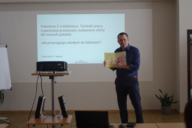 mężczyzna w siwej koszule i czarnych spodnia trzyma w dłoniach książkę, w tle ekran z napisami, po lewej stronie rzutnik