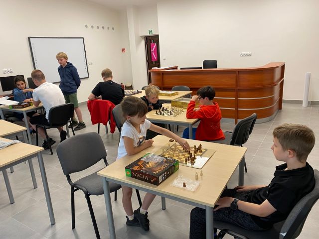 przy brązowych stołach siedzą dzieci i grają w szachy, przed nimi stoi mężczyzna