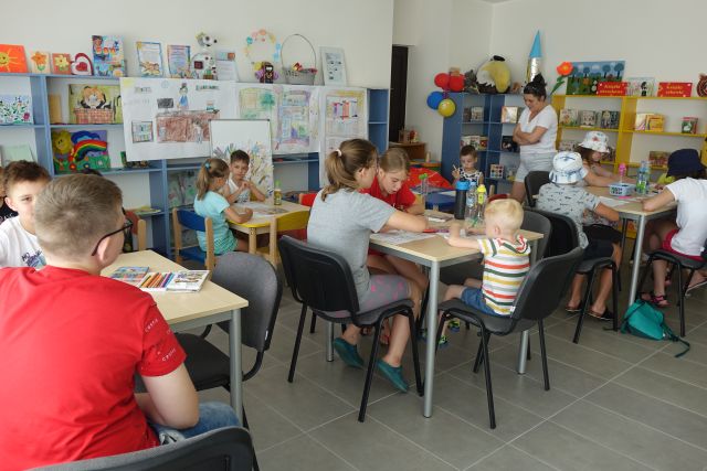 przy brązowych stołach siedzą dzieci i wypełniaja karty pracy, za nimi niebieskie regały