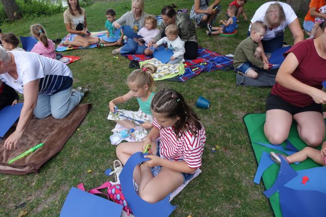 dzieci i opiekunowie siedzą na trawie, dzieci wycinają z niebieskiego papieru łuk